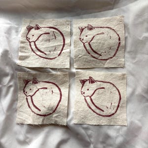 May include: Four square fabric patches with a burgundy cat illustration. The cat is curled up in a sleeping position. The patches are made of a natural-colored fabric and are arranged in a square.