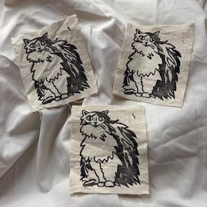 May include: Three square patches with a black and white illustration of a fluffy cat. The cat is sitting upright with a detailed outline and a textured, shaggy appearance. The patches are made of a light-colored fabric.