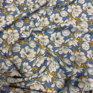 May include: Blue fabric featuring a floral pattern of large white and pale blue flowers with yellow details. The fabric's creases indicate it's a soft material, possibly cotton or linen.