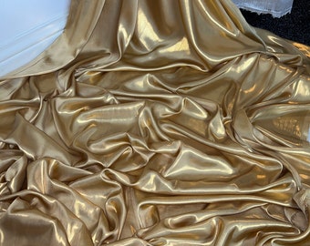 Op de afbeelding: Een gouden metallic stof met een gladde, zijdeachtige textuur. De stof is gedrapeerd en gevouwen, wat een luxe en elegante uitstraling creëert.