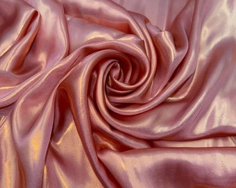 Op de afbeelding: Close-up van een zachte, roze satijnen stof met een glinsterende, iriserende afwerking. De stof is gedrapeerd en gevouwen, wat een luxe en elegante uitstraling creëert.