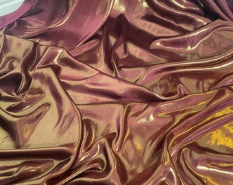Op de afbeelding: Een close-up van een bordeauxrode stof met een gouden metallic glans. De stof is gedrapeerd en gevouwen, waardoor zachte, vloeiende lijnen ontstaan.
