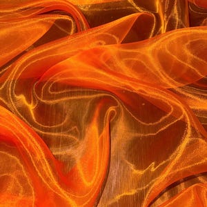 Puede incluir: Una tela de organza de color naranja vibrante que presenta una textura transparente y ligera con un tacto rígido. El material está drapeado en pliegues suaves y fluidos que crean variadas profundidades de color y reflejos de luz.
