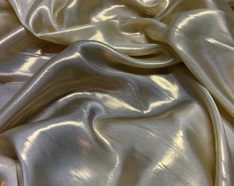 Op de afbeelding: Close-up van een glinsterende, vloeiende stof in een zachte, crèmekleurige gele tint. Het materiaal heeft een glanzende glans, met highlights en schaduwen die een gevoel van diepte en beweging creëren. De stof lijkt gedrapeerd te zijn, met zachte plooien en rondingen.