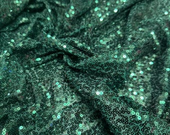 May include: Emerald green fabric with sequins. The sequins are small and round, and they are densely packed together. The fabric is wrinkled and has a soft, luxurious feel.