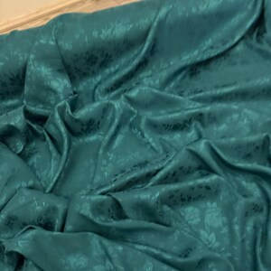 May include: Teal-colored fabric with a floral pattern. The fabric is draped, showing folds and shadows that highlight the texture and design. The sheen suggests a satin or silk material.