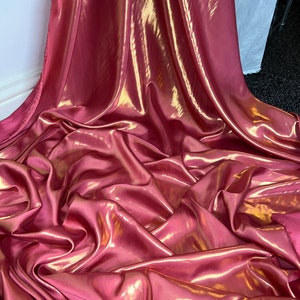 Op de afbeelding: Close-up van een gedrapeerde stof met een glanzende, iriserende afwerking. De stof heeft een diepe roze kleur met gouden highlights, waardoor een glinsterend effect ontstaat. De plooien en rondingen van het materiaal geven diepte en textuur aan de afbeelding.
