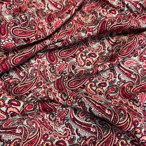 May include: Close-up of a fabric with a repeating paisley pattern. The design features intricate, swirling shapes in shades of red, burgundy, and peach against a light blue background. The fabric appears soft and flowing.