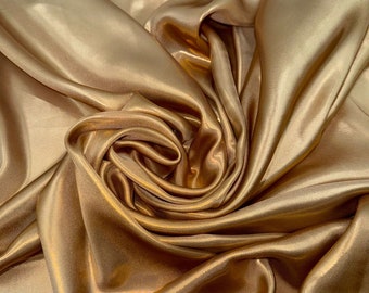 Op de afbeelding: Een close-up van een gouden satijnen stof met zachte, vloeiende plooien en een luxe glans.