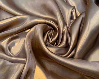 Op de afbeelding: Close-up van een gladde, gouden satijnen stof met een subtiele glans. De stof is gedrapeerd en gevouwen, waardoor zachte, vloeiende curves ontstaan.