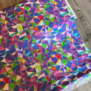 Puede incluir: Una tela vibrante con un patrón geométrico de triángulos coloridos en tonos morado, azul, verde, rosa y amarillo. La tela está doblada, revelando un borde blanco, y descansa sobre una superficie de madera.