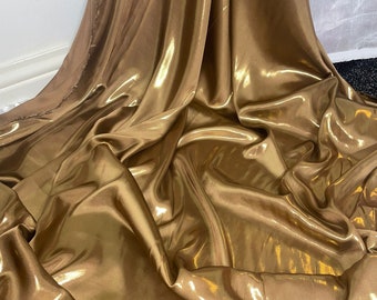 Op de afbeelding: Een close-up van een gouden metallic stof met een glad, glanzend oppervlak. De stof is gedrapeerd en gevouwen, waardoor zachte, vloeiende lijnen ontstaan.