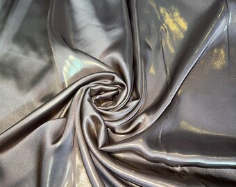 Op de afbeelding: Close-up van een gladde, iriserende stof met een metallic glans. De stof is gedrapeerd en gevouwen, waardoor zachte, vloeiende lijnen ontstaan.