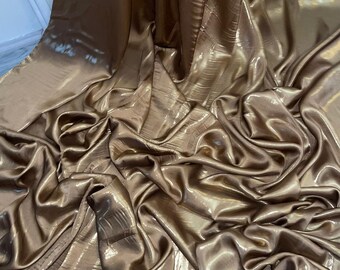 Op de afbeelding: Een close-up van een metallic gouden stof met een subtiele glans. De stof is gedrapeerd en gevouwen, waardoor de zachte textuur zichtbaar wordt.