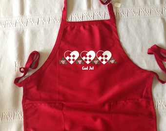 May include: Red Christmas apron with three white hearts and the text "God Jul" in white. The apron has two pockets.