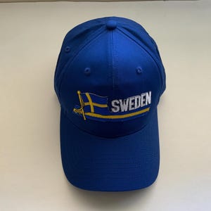 May include: A royal blue baseball cap featuring the word "SWEDEN" in white lettering, with a yellow and blue flag design. The cap has a curved brim and a button on top.