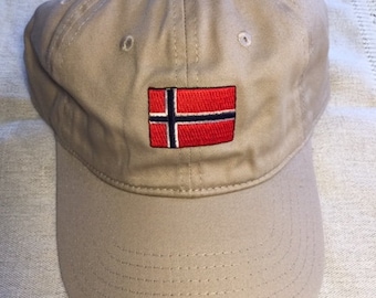 May include: A tan baseball cap with a curved brim. The cap features a rectangular patch with the flag of Norway, which is red with a white-bordered blue cross. The cap is made of a durable fabric and has a button on top.