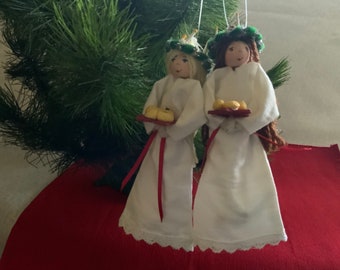 May include: Two white fabric dolls with long brown hair wearing white gowns and green wreaths on their heads. They are holding trays of small yellow breads.
