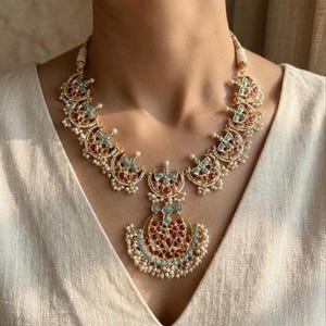 May include: A gold-toned necklace featuring a series of crescent-shaped links adorned with inlaid red and light blue stones. The central pendant is a larger crescent shape with matching stones and small white pearl-like beads along the bottom edge.