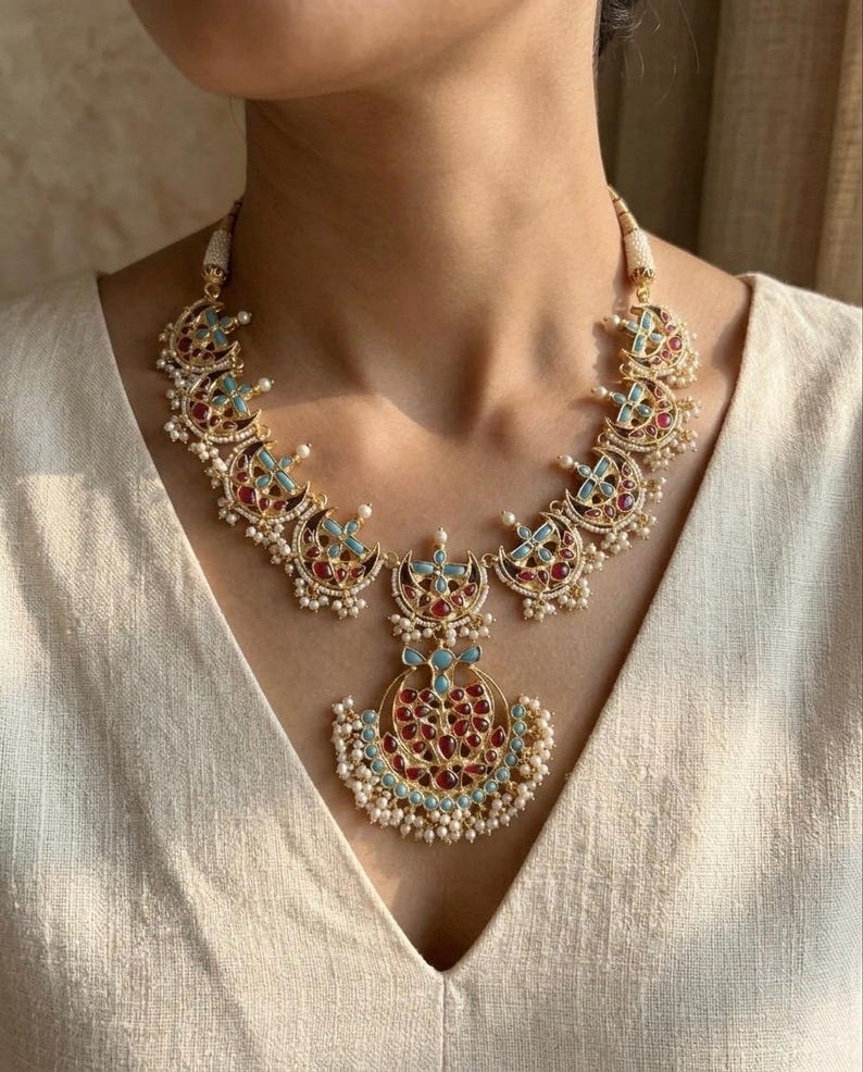 May include: A gold-toned necklace featuring a series of crescent-shaped links adorned with inlaid red and light blue stones. The central pendant is a larger crescent shape with matching stones and small white pearl-like beads along the bottom edge.