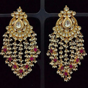 May include: A pair of gold-toned chandelier earrings. Each earring features a large, teardrop-shaped clear stone at the center, surrounded by smaller clear stones. White pearls and ruby-colored stones dangle from the bottom
