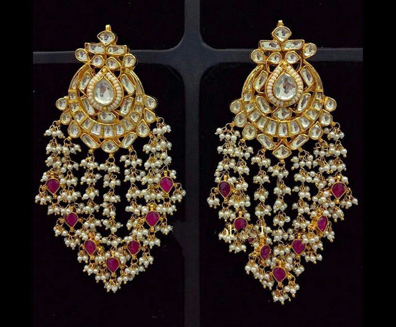 May include: A pair of gold-toned chandelier earrings. Each earring features a large, teardrop-shaped clear stone at the center, surrounded by smaller clear stones. White pearls and ruby-colored stones dangle from the bottom