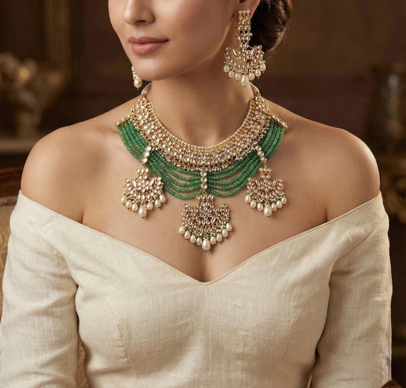 May include: A gold and green Indian bridal necklace set with pearl and emerald accents. The necklace features a layered design with a large, ornate pendant. The earrings are similarly designed with a smaller pendant