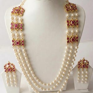 Puede incluir: Un collar de perlas de tres hilos con pendientes a juego. El collar presenta detalles dorados y engastes de piedras rojas cuadradas. Los pendientes tienen un diseño similar, con hilos de perlas colgantes.
