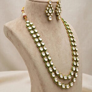 May include: A gold-toned necklace and earring set featuring teardrop-shaped white stones outlined in green enamel. The necklace consists of two long strands of connected stones, while the matching earrings are smaller, elongated teardrop shapes.