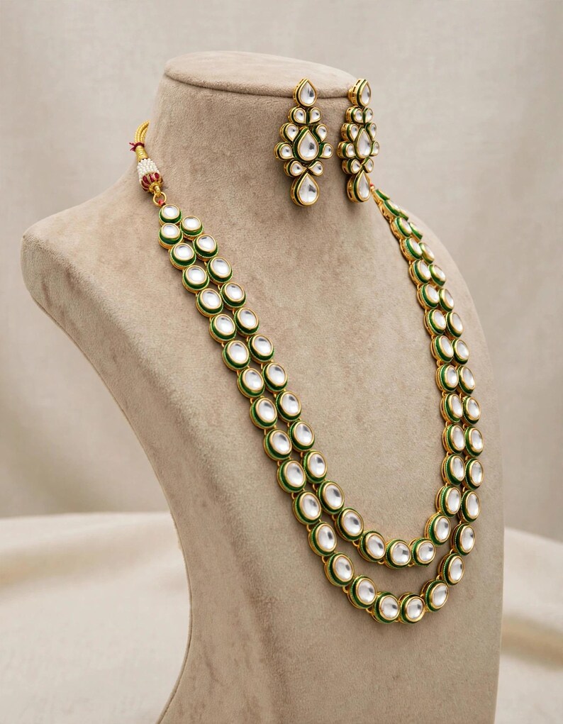 May include: A gold-toned necklace and earring set featuring teardrop-shaped white stones outlined in green enamel. The necklace consists of two long strands of connected stones, while the matching earrings are smaller, elongated teardrop shapes.