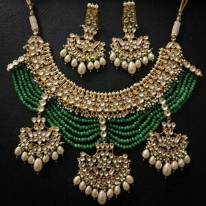 May include: A gold and green Indian bridal necklace set with pearl and emerald accents. The necklace features a layered design with a large, ornate pendant. The earrings are similarly designed with a smaller pendant