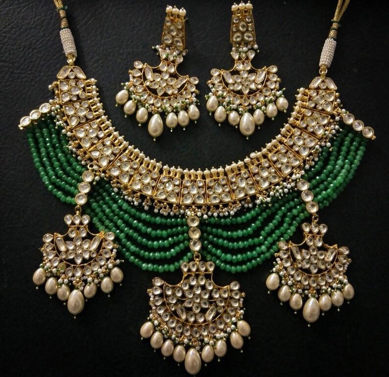 May include: A gold and green Indian bridal necklace set with pearl and emerald accents. The necklace features a layered design with a large, ornate pendant. The earrings are similarly designed with a smaller pendant