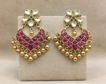 May include: A pair of gold-toned earrings featuring a crescent-shaped drop design inlaid with deep pink stones in a grid pattern. The top section is a floral motif with white stones and small green accents, finished with dangling gold beads.