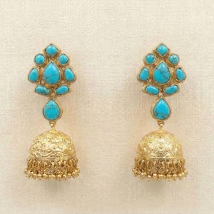 May include: A pair of gold-colored earrings with turquoise-colored stones. The earrings have a floral design on the bell-shaped bottom and a cluster of turquoise stones at the top. The earrings are displayed on a cream-colored background.