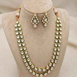 May include: A two-strand gold-tone necklace featuring round white stones set in green enamel borders, accompanied by a pair of matching drop earrings with a cluster of white stones in a diamond-shaped arrangement with green enamel detailing.