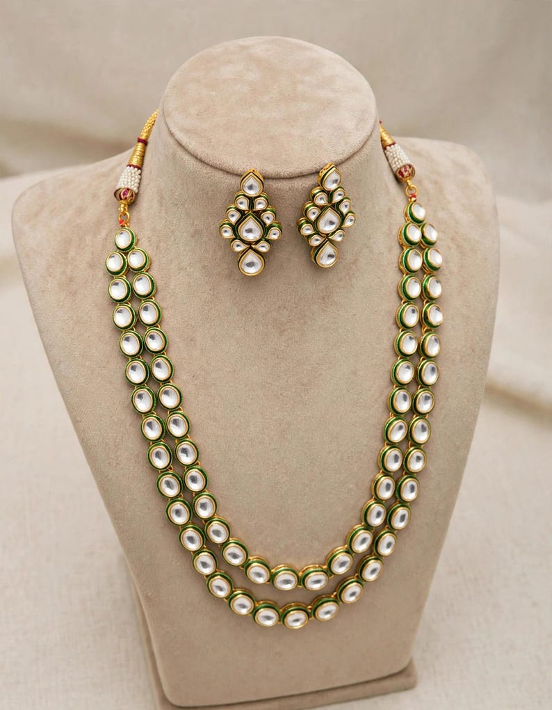 May include: A two-strand gold-tone necklace featuring round white stones set in green enamel borders, accompanied by a pair of matching drop earrings with a cluster of white stones in a diamond-shaped arrangement with green enamel detailing.