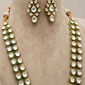 May include: A jewellery set consisting of a double-strand necklace and matching drop earrings. Each piece features oval and teardrop-shaped white stones set in gold-toned metal with green enamel borders. The necklace includes small white pearl accents.