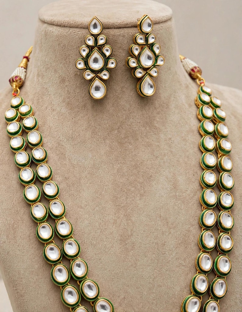 May include: A jewellery set consisting of a double-strand necklace and matching drop earrings. Each piece features oval and teardrop-shaped white stones set in gold-toned metal with green enamel borders. The necklace includes small white pearl accents.