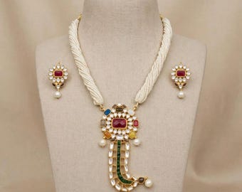May include: A necklace and earring set featuring a multi-strand white seed bead chain. The central pendant is gold-toned with a large rectangular red stone, surrounded by smaller colourful stones and a curved drop section with green stones and pearls.
