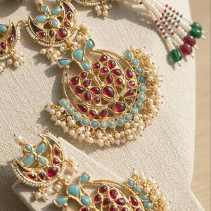 May include: A gold-toned necklace and earring set featuring crescent-shaped pendants adorned with red and turquoise stones. The jewelry is embellished with small white seed pearls and includes a gold cord with beaded tassels at the ends.