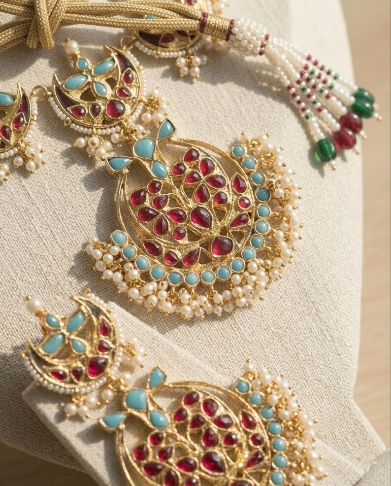 May include: A gold-toned necklace and earring set featuring crescent-shaped pendants adorned with red and turquoise stones. The jewelry is embellished with small white seed pearls and includes a gold cord with beaded tassels at the ends.