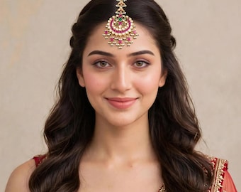 May include: A gold maang tikka headpiece featuring a crescent-shaped design adorned with small white pearls, vibrant pink stones, and a central green stone. The jewelry piece is centered on the forehead, showcasing intricate metalwork and dangling beads.