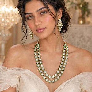 May include: A two-strand necklace featuring oval white stones set in gold-toned metal with green enamel borders, paired with matching drop earrings. Each earring consists of a cluster of oval white stones with green enamel detailing.