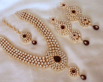 May include: A traditional Indian bridal jewelry set featuring a gold necklace, earrings, and a maang tikka. The set is adorned with white and clear stones and red accents.