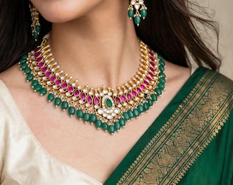 May include: A gold-toned necklace and matching drop earrings set featuring teardrop-shaped fuchsia stones, round green beads, and clear white accents. The necklace has a layered design with a central pendant and dangling green bead fringe.