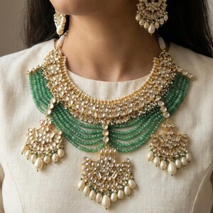 May include: A gold and green Indian bridal necklace set with pearl and emerald accents. The necklace features a layered design with a large, ornate pendant. The earrings are similarly designed with a smaller pendant
