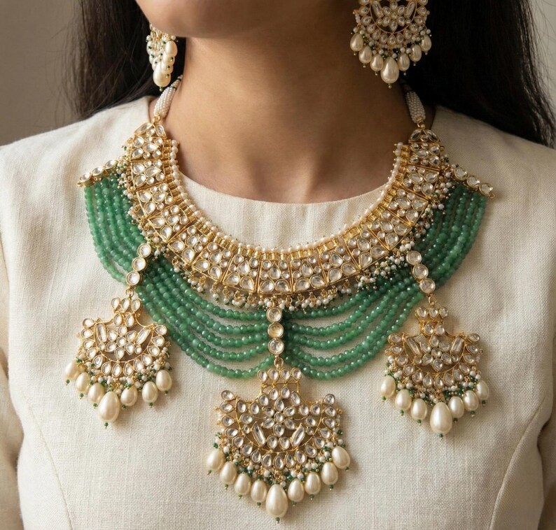 May include: A gold and green Indian bridal necklace set with pearl and emerald accents. The necklace features a layered design with a large, ornate pendant. The earrings are similarly designed with a smaller pendant