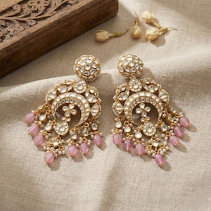 Peut inclure: Une paire de boucles d'oreilles pendantes dorées avec des détails complexes en pierres blanches et transparentes. Les boucles d'oreilles présentent une forme de croissant avec des perles roses pendantes et de petites perles blanches. Elles sont posées sur un tissu neutre.
