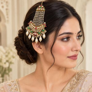 May include: A gold-toned hair accessory featuring a crescent-shaped base adorned with red and green stones, accented by multiple strands of small white beads and larger teardrop-shaped pearl-like pendants hanging from the bottom edge.