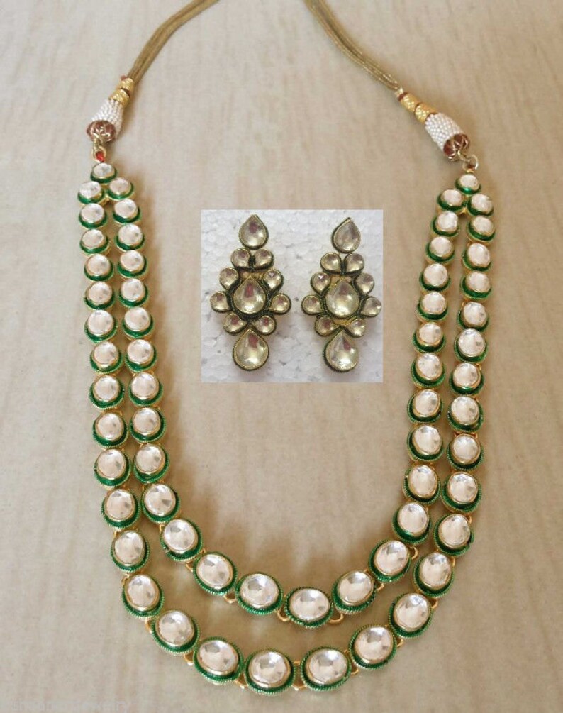 May include: A two-strand necklace featuring round, clear stones set in green metal frames, connected to a gold-toned cord with pearl-like accents. Included are two matching drop earrings with a cluster of clear stones in green metal settings.